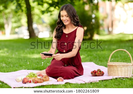 leisure and people concept - happy smiling woman with picnic basket and smartphone photographing food on blanket at summer park