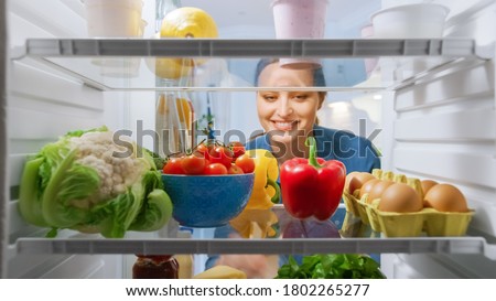 Beautiful Young Woman Looks inside the Fridge and Takes out Vegetables. Woman Preparing Healthy Meal Using Groceries. Point of View POV from Inside of the Kitchen Refrigerator full of Healthy Food Royalty-Free Stock Photo #1802265277