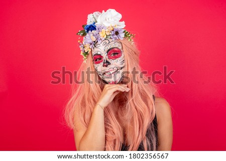 Lovely woman wears halloween makeup, dressed in black outfit, red wreath, has zombie image Optimistic keeps hands partly crossed and hand under chin, looks at camera with pleasure. Happy emotions