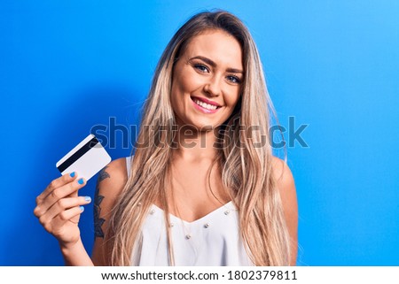 Young beautiful blonde woman holding credit card over isolated blue background looking positive and happy standing and smiling with a confident smile showing teeth