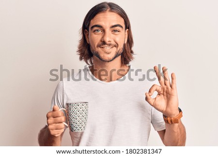 Young handsome man holding coffee doing ok sign with fingers, smiling friendly gesturing excellent symbol 