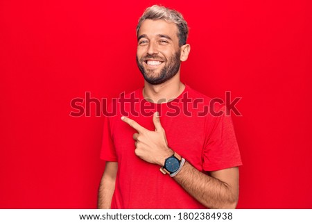 Young handsome blond man wearing casual red t-shirt standing over isolated red background smiling cheerful pointing with hand and finger up to the side