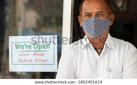 Small business owner in medical mask standing in front of door with we are open wear mask notice board - concept of support local community and buy local during coronavirus or covid-19.