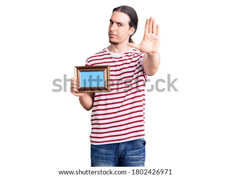 Young adult man with long hair holding empty frame with open hand doing stop sign with serious and confident expression, defense gesture 