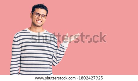 Hispanic handsome young man wearing casual clothes and glasses smiling cheerful presenting and pointing with palm of hand looking at the camera. 