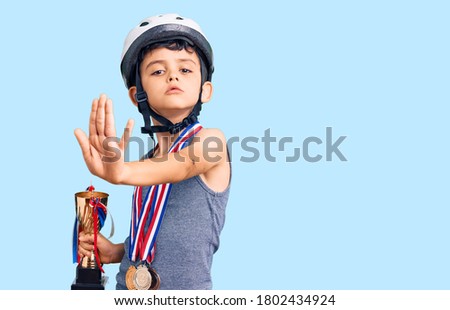 Little cute boy kid wearing bike helmet and winner medals holding winner trophy with open hand doing stop sign with serious and confident expression, defense gesture 