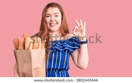 Young beautiful redhead woman holding paper bag with bread doing ok sign with fingers, smiling friendly gesturing excellent symbol 