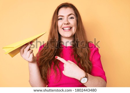 Young redhead woman holding paper airplane smiling happy pointing with hand and finger 
