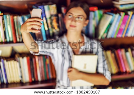 Young beautiful brunette in dress and with eyeglasses holding a book and taking selfie while standing in library. Selective focus on hand.