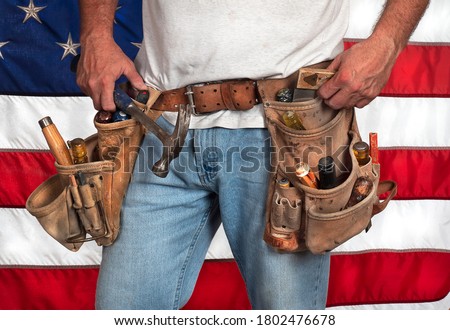Closeup carpenter wearing worn out old, leather toolbelt with hand tools in front of American flag, construction, american pride, building, made in america, labor, laborer, worker, tradesman Royalty-Free Stock Photo #1802476678