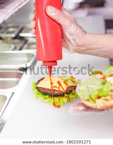 Cook adding a ketchup on burger. Preparing burger in restaurant. Hands of the cook in disposable gloves based on hygiene requirements.