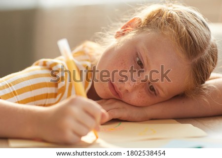 Warm toned portrait of cute red haired girl drawing pictures while lying her head on table in sunlight