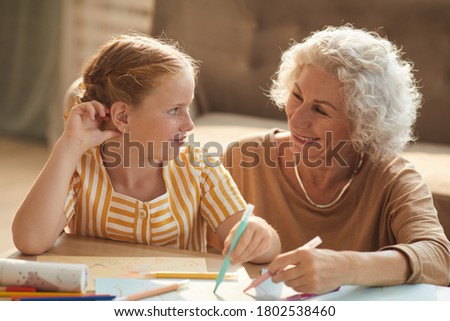 Warm toned portrait of smiling senior woman looking at cute red haired girl and drawing together while sitting on floor by coffee table in cozy living room.