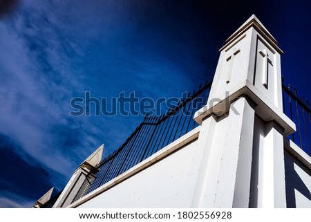 High white concrete walls with metal bars separate the dead from an outside cemetery, background with a blue sky.