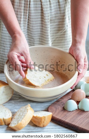 Woman's hands dipping bread into French toast batter with farm fresh organic eggs and a loaf of bread nearby.