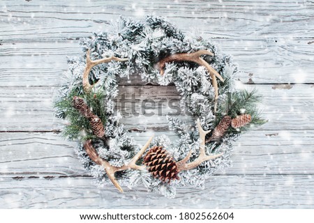 Homemade flocked Christmas hunter's wreath over a white rustic wooden background with falling snow. Top view or flatlay position.