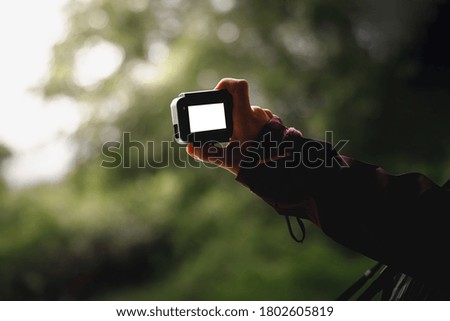 Hand holding action camera with white display screen and blur green forest background.
