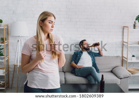 Latent alcoholism. Sad wife sees her husband drinking a lot of beer, man with beard sits on couch, near table with empty bottles Royalty-Free Stock Photo #1802609389