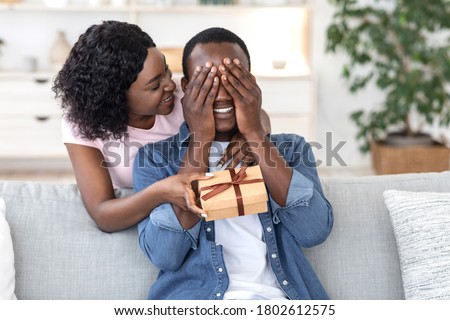Black woman giving her boyfriend birthday gift at home, man covering his eyes with palms, copy space