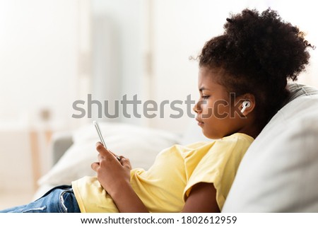 African Teenager Girl Using Smartphone And Earbuds Listening To Educational Podcast Sitting On Couch At Home. Applications For Teens Concept