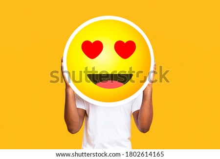 Unrecognizable black man hiding face against in love emoticon emoji, holding big illustrated smile sticker over yellow background, copy space Royalty-Free Stock Photo #1802614165