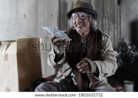 Poor homeless old man smiling and holding face mask while sitting in the corner of the building near the wall blur face