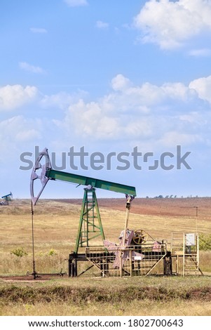 Oil pump jack in operation on agricultural field, blue sky, vertical