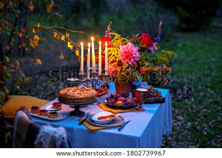 Autumn evening photo shoot - romantic dinner outdoors. Table with tablecloth and decoration - pie, figs, glasses, plates, table setting and candelabra with candles. Fall flowers dahlia bouquet.