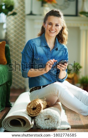 smiling modern middle age woman in jeans shirt and white pants with pile of carpets searching for carpet cleaning service on a smartphone in the modern house in sunny day.