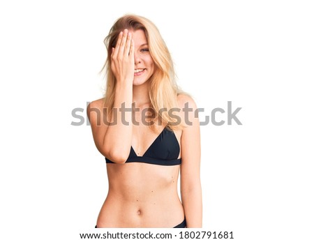 Young beautiful blonde woman wearing bikini covering one eye with hand, confident smile on face and surprise emotion. 