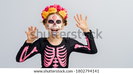Adorable zombie in flower wreath posing on grey background. Happy child with Halloween creative makeup. Girl celebrating for Mexican Day of the Dead.