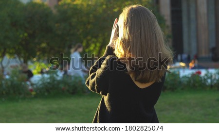 Rear view of woman in black sweater holding photocamera and taking photo at summer green park. Media. Young girl photographing sights in a big city in the summertime.