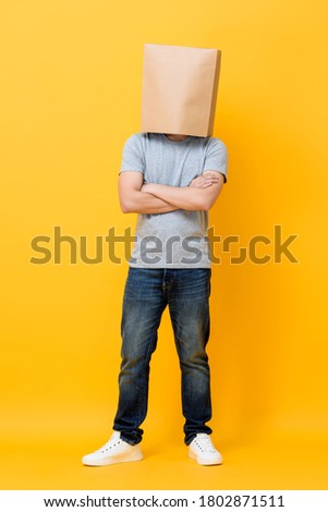 Concept full length portrait of Anonymous man with head covered with paper bag doing arms crossed gesture in yellow studio background Royalty-Free Stock Photo #1802871511