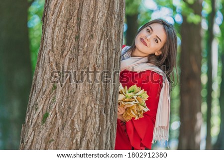 Winsome caucasian woman with a bouquet of leaves looks from behind a tree outdoors in autumn park.
