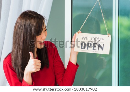 Asian young woman smile show finger thumb up for good sign she notice sign wood board label "WELCOME OPEN" hang through glass door front shop, Business turning open after coronavirus pandemic disease