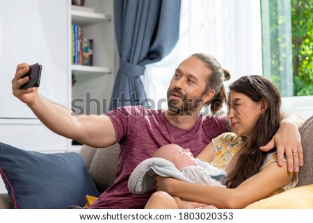 Caucasian Man using smartphone selfie with his wife and sleeping newborn baby son. Father and mother spending time together with cute infant child boy at home. Happy Family and baby healthcare concept