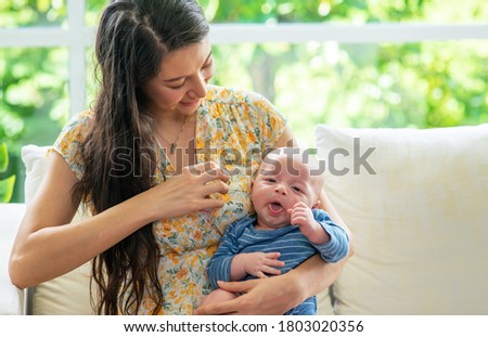 Smiling Caucasian woman mother holding and playing with sleepy newborn baby son. Beautiful mom sitting on sofa with embracing her infant child boy. Happy family, maternity and baby healthcare concept.