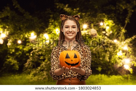 holiday, theme party and people concept - happy smiling woman in halloween costume of leopard with ears holding jack-o-lantern pumpkin over festive garland lights at night park on background