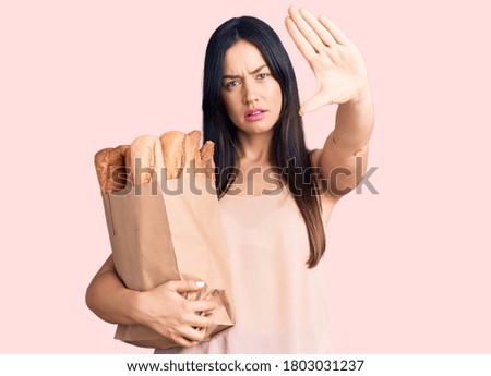 Young beautiful caucasian woman holding paper bag with bread with open hand doing stop sign with serious and confident expression, defense gesture 