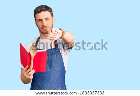 Handsome young man with bear wearing professional baker apron reading cooking recipe book with angry face, negative sign showing dislike with thumbs down, rejection concept 
