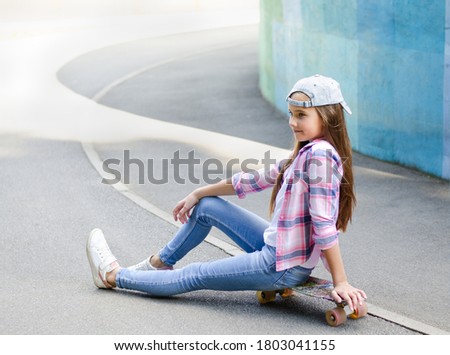 Smiling cute little girl child in cap sitting on a skateboard. Preteen with penny board outdoors in summer day
