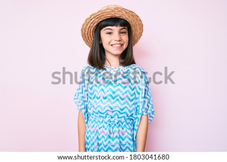 Young little girl with bang wearing summer dress and hat looking positive and happy standing and smiling with a confident smile showing teeth 