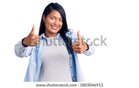 Hispanic woman with long hair wearing casual denim jacket approving doing positive gesture with hand, thumbs up smiling and happy for success. winner gesture. 