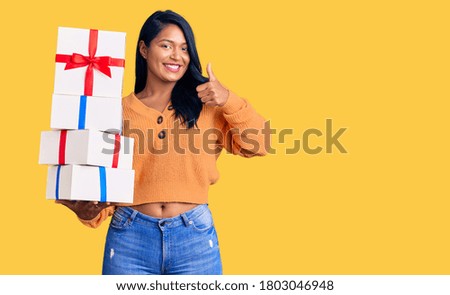 Hispanic woman with long hair holding gifts smiling happy and positive, thumb up doing excellent and approval sign 
