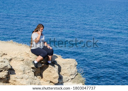 a young, beautiful woman speaks by phone, sitting on a cliff. Against the background of the sea