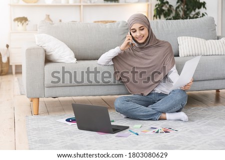 Smiling arabic female graphic designer working from home, discussing project with client on cellphone and using laptop, checking swatches, free space