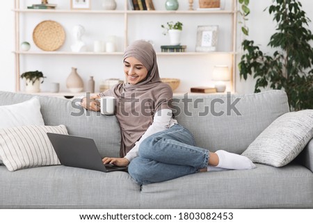 Positive arabic woman unwinding with laptop and coffee on couch at home, relaxing in stylish living room Royalty-Free Stock Photo #1803082453