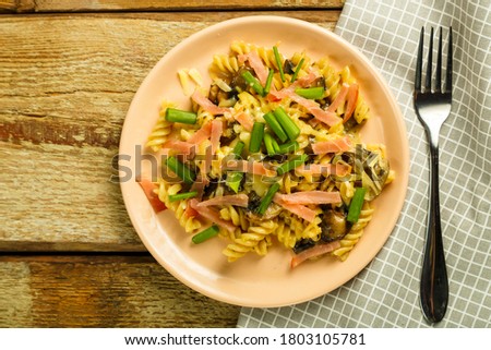 a plate of pasta with ham and mushrooms in a creamy sauce on the table with a fork on a napkin.