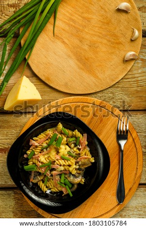 Black plate with pasta with ham and mushrooms in a creamy sauce on the table with a fork near the ingredients.