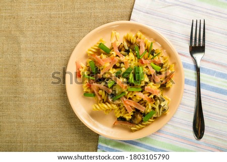 a plate of pasta with ham and mushrooms in a creamy sauce on the table with a fork on a napkin on the tablecloth.